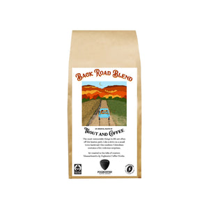 A brown bag labeled "Back Road Blend" by Trout & Coffee shows a blue truck on a dirt road, colorful hills, and a sunset sky, highlighting this fresh roasted, medium roast original coffee blend.
