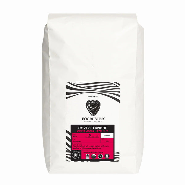 A white bag of Fogbuster® Coffee Works Covered Bridge ground coffee features black and white patterns, a strawberry icon, and pink accents. It’s a medium roast organic coffee with cinnamon and hazelnut notes.