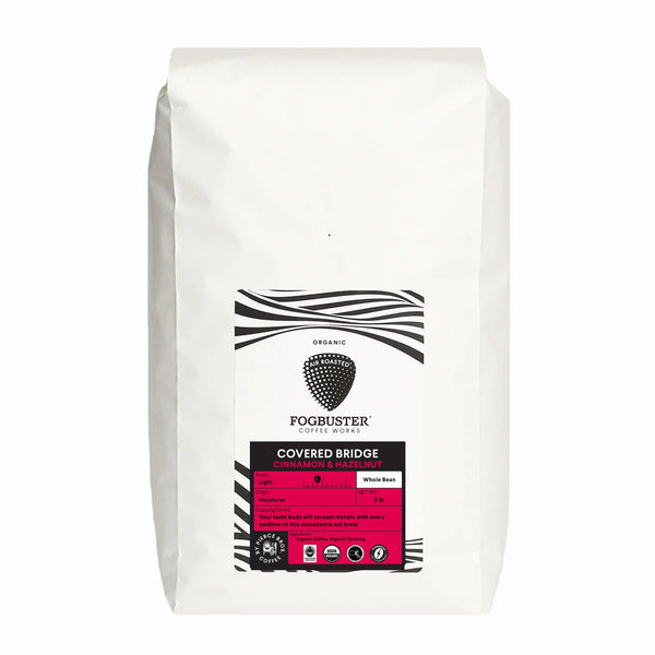 A 907g white bag of Fogbuster® Coffee Works’ "Covered Bridge" light roast features whole beans from Honduras with aromatic cinnamon and hazelnut notes. The label is mainly white and black with pink accents.