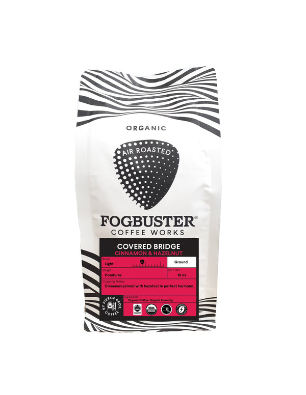 A white-and-black striped bag of Fogbuster® Coffee Works Covered Bridge Honduras ground coffee, with cinnamon and hazelnut notes on a pink label; organically grown and air roasted.