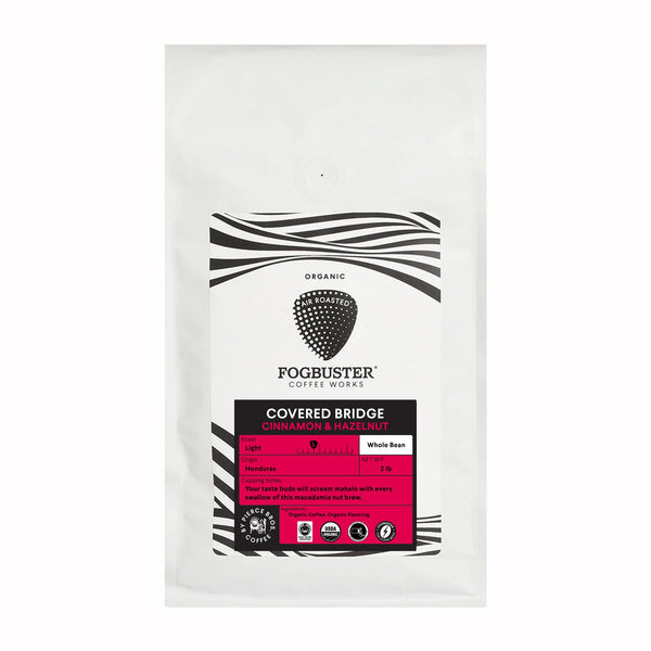 A 2 lb white bag of Fogbuster® Coffee Works whole bean coffee, "Covered Bridge," features black and pink accents, Honduras beans, and notes of cinnamon and hazelnut.