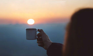 A person holds up a white mug against the backdrop of a serene sunrise over a foggy landscape. The sun hovers just above the horizon, casting a warm, orange glow across the scene. The atmosphere is tranquil and refreshing.