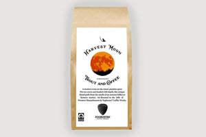 A brown paper coffee bag labeled "Harvest Moon" from Trout & Coffee, featuring an orange moon above black tree silhouettes. The label describes the fresh roasted coffee blend and displays the Fogbuster Coffee Works logo at the bottom.