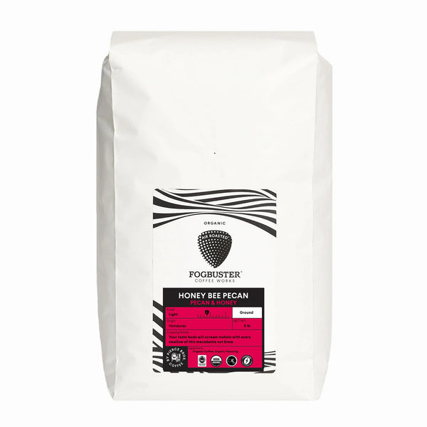 A white bag of Fogbuster® Coffee Works Honey Bee Pecan features a black and white label with a honeycomb logo and pink accents, highlighting its light roast flavor and roast details.