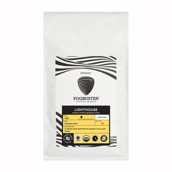 A white bag of Fogbuster® Coffee Works Lighthouse whole bean light roast coffee features a black and white label, caramel notes, organic certification, fair trade and compostable packaging icons.