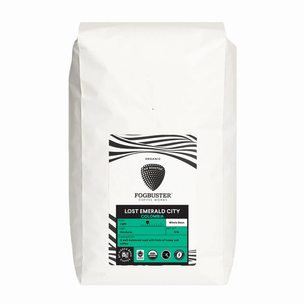 A white bag of Fogbuster® Coffee Works Lost Emerald City coffee, featuring black and white graphics with a turquoise accent, offers organic Colombian beans with tasting notes of honey, red fruit, and citrus almond.