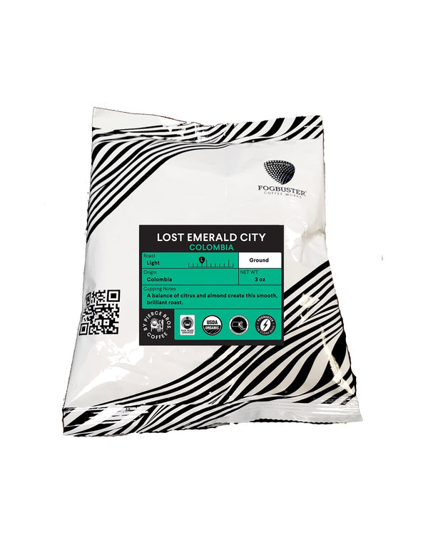 A white bag with black zebra stripes labeled "Lost Emerald City" by Fogbuster® Coffee Works. Teal label notes this Colombian light roast, ground, 12 oz, features vibrant citrus and almond flavors.