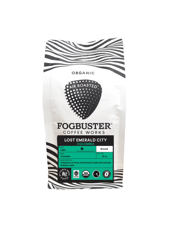 A white bag of Fogbuster® Coffee Works "Lost Emerald City" light roast coffee with citrus almond notes, black zebra stripes, and icons for organic, air roasted, and ground Colombian coffee.