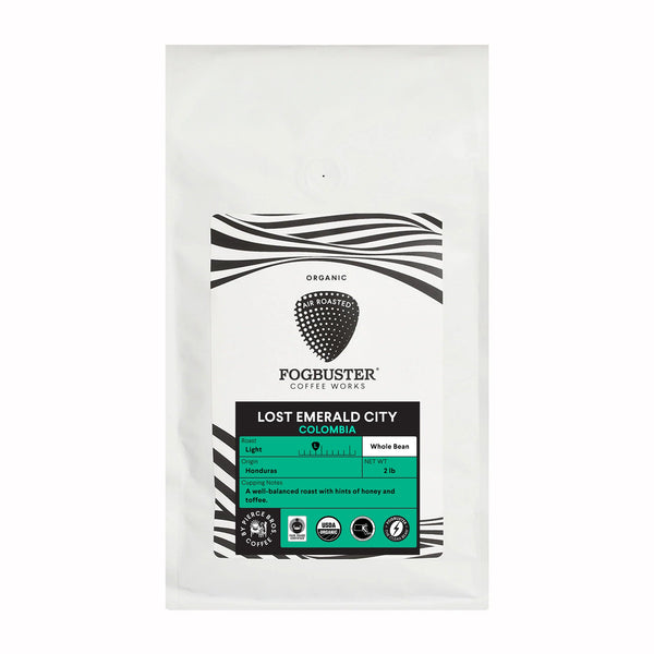 A white bag of Fogbuster® Coffee Works Lost Emerald City coffee displays a black logo, wavy lines, and a green label describing it as organic, medium roast with honey, citrus almond, and coffee notes.