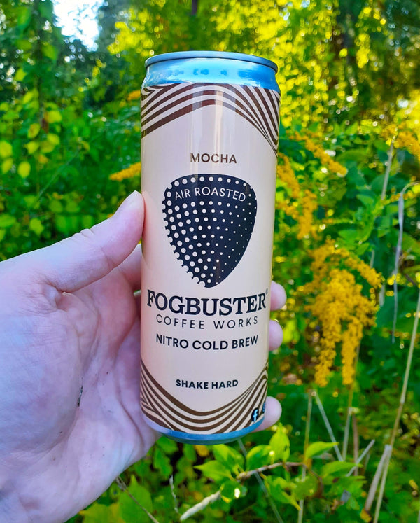 A hand holds a can of Mocha by Fogbuster® Coffee Works in front of green leaves and yellow flowers.