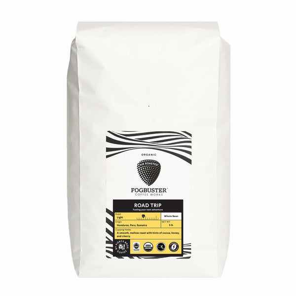 A white bag of Fogbuster® Coffee Works "Road Trip" organic whole bean coffee, with cocoa, honey, and berry notes, features a minimalist black and white design accented with yellow on the label.