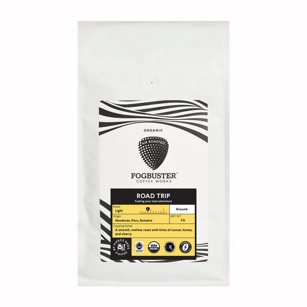 Fogbuster® Coffee Works "Road Trip" light roast comes in a white bag with black and white wavy lines. This Honduras coffee is smooth and mellow, with cocoa, honey, and citrus notes. Organic and Fair Trade icons are displayed.