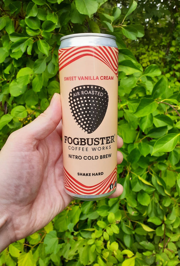 A hand holds a can of Fogbuster® Coffee Works Sweet Vanilla Cream Nitro Cold Brew against green leafy bushes. The shelf-stable can instructs to "shake hard" before drinking.