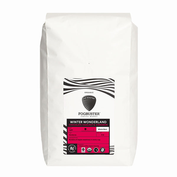 A white bag of Fogbuster® Coffee Works Winter Wonderland whole bean coffee features a black and white label with pink accents and notes of chocolate, roasted hazelnuts, and cinnamon from Honduras.