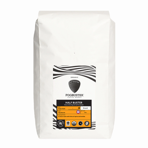 A white Fogbuster® Coffee Works bag labeled “Half Buster,” featuring a black coffee bean, orange stout roast mark, and displaying its half decaf ground coffee blend in a 1 lb size.