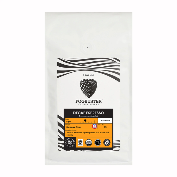 A white bag of Fogbuster® Coffee Works Decaf Espresso displays a black logo, orange label, and highlights its organic whole bean Honduras Timor coffee blend.