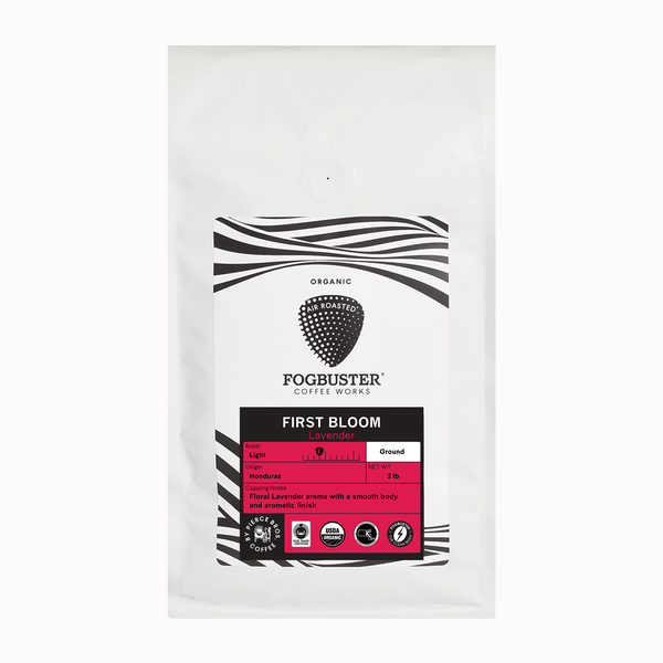 A white bag of Fogbuster® Coffee Works First Bloom, a light roast with floral lavender notes, has a black and white label with a raspberry graphic and product details such as origin, roast level, and tasting notes.