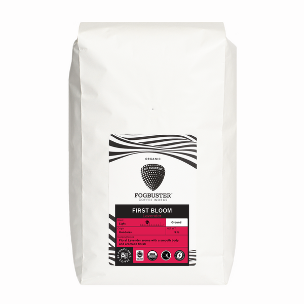 A white Fogbuster® Coffee Works bag labeled "First Bloom" features organic ground coffee with floral lavender flavor, a light roast, black and white patterns, and a pink section indicating roast level and weight.
