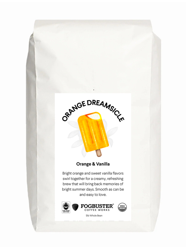 A white coffee bag labeled "Orange Dreamsicle" from Fogbuster® Coffee Works features an orange popsicle illustration. The label describes the light roast as having orange and vanilla flavors.