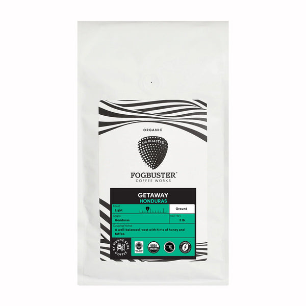 A white coffee bag labeled "Fogbuster® Coffee Works" with a black and white pattern features a green label that reads "Fundraiser - Getaway (Single Origin - Honduras coffee)." This light roast, 2 lbs coffee has tasting notes of honey and citrus. It is organic, ground, and air-roasted.