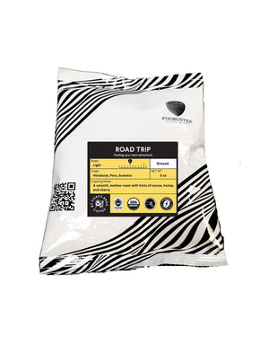 A white and black patterned coffee bag labeled "Road Trip" by Fogbuster® Coffee Works. This Honduras ground coffee is a smooth, mellow light roast (12 oz) with cocoa, honey, and cherry notes. USDA Organic and Fair Trade certified.