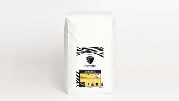 A white coffee bag labeled "Air-Roasted - Single Serve K-Cups" with a black and yellow design. The label reads "Fogbuster® Coffee Works" and indicates the coffee is organic, 12oz in weight, and 340 grams. The packaging features waves and dot patterns, highlighting the premium shade-grown coffee beans used.