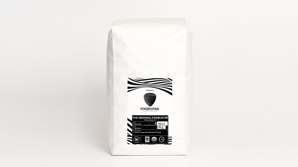 A white coffee bag with a black and white label displaying "Fogbuster® Coffee Works" at the top. The label design features a striped pattern and text indicating "THE ORIGINAL FOGBUSTER," complemented by certifications for organic and fair trade. Made from premium shade-grown coffee utilizing advanced air roasting technology, this gourmet blend is available in low acid, single-serve K-Cups.