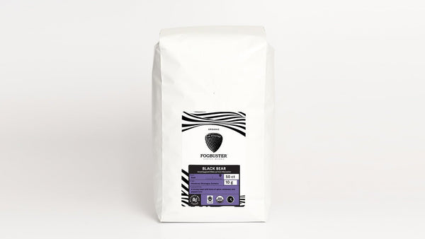 A white bag of coffee from Fogbuster® Coffee Works, featuring a black logo with a stylized coffee bean design. The label reads "Air-Roasted - Single Serve K-Cups" and "Black Bear," along with additional information about the product's origin and roast level. Enhanced through air roasting technology, there's also a lavender-colored section on the lower part of the label.