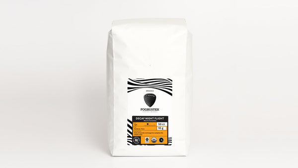 A white coffee bag with a label that reads "Fogbuster® Coffee Works" and "Air-Roasted - Single Serve K-Cups (Gourmet - Low Acid - Single Serve Coffee)" in the center. The label features a black and white abstract design at the top, an orange section at the bottom detailing its premium shade-grown origins, and highlights its roast profile enhanced by air roasting technology.