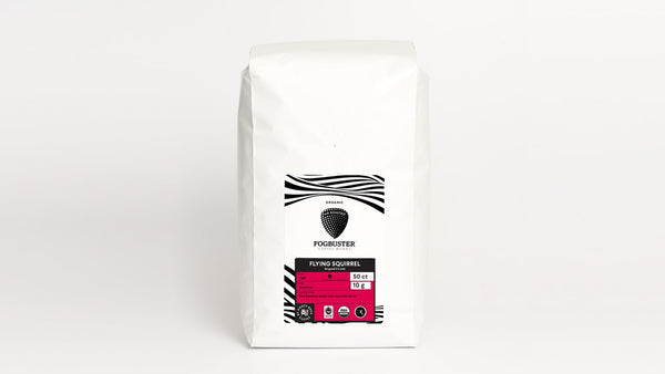 A white bag of Air-Roasted, Single Serve K-Cups from Fogbuster® Coffee Works, labeled "Flying Squirrel," rests against a plain background. The label features a black and pink design, indicating the premium shade-grown coffee is organic and low-acid, weighing 10 ounces.