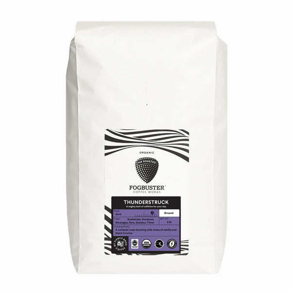 A white coffee bag displays the label "Fogbuster® Coffee Works - Thunderstruck." The design includes black and white stripes, with a purple section highlighting its French roast ground coffee featuring high caffeine content. With tasting notes of vanilla and chocolate, this premium shade-grown blend offers a delightful experience.