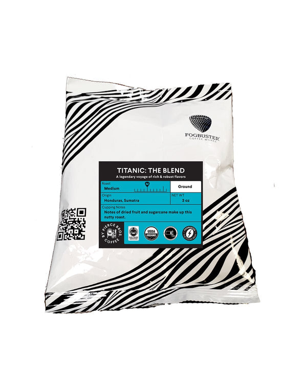 A 12 oz white package of "Titanic: The Blend" by Fogbuster® Coffee Works, showcasing a black and white striped design with a blue label, contains Organic Fair Trade medium roast coffee from Honduras and Sumatra. The package also includes a QR code.