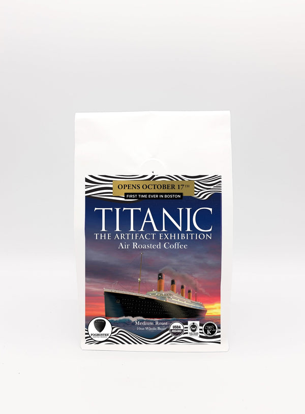 A white coffee bag features an image of the Titanic, adorned with the text "Titanic: The Blend," by Fogbuster® Coffee Works. This air-roasted, medium roast coffee opens on October 17th and is noted for its Organic, Fair Trade, and Kosher origins. Logos and additional details further emphasize these qualities.